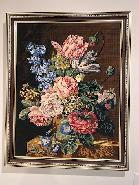 A framed needlepoint or tapestry featuring a detailed floral bouquet with pink, red, blue, and white flowers on a dark brown background, enclosed in a decorative silver-toned frame and displayed on a light wall.